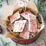 A circular tin lined with brown paper filled with homemade peppermint bark, surrounded by holiday garland and more pieces of bark.