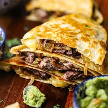 A stack of three freshly prepared fajita steak quesadillas sitting on a wooden cutting board next to a small bowl filled with homemade guacamole.