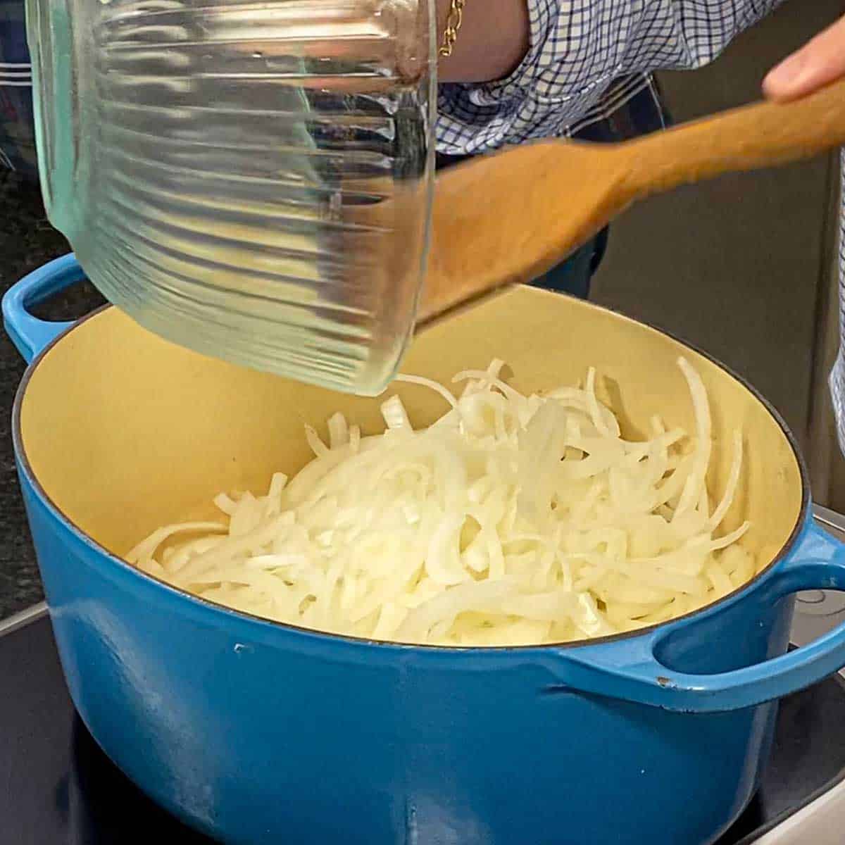 A person using a large wooden spatula to transfer thinly sliced onions from a glass bowl into a large oval Dutch oven sitting on an induction stovetop.