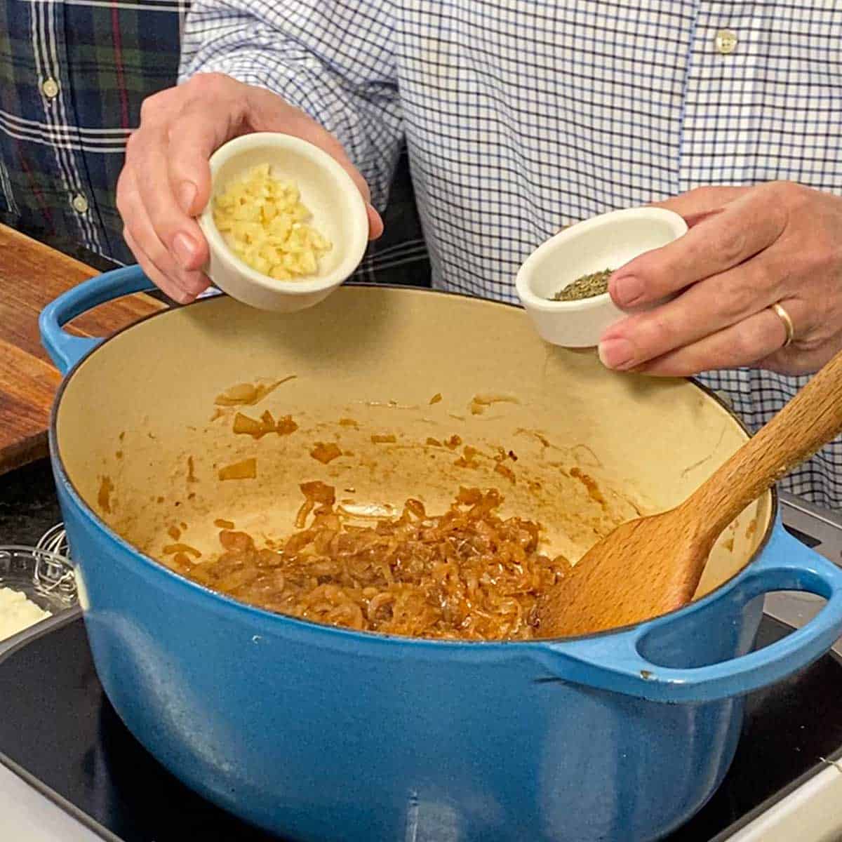 A person getting ready to dump minced garlic from one bowl and dried thyme from another small bowl into a large Dutch oven filled with caramelized onions.