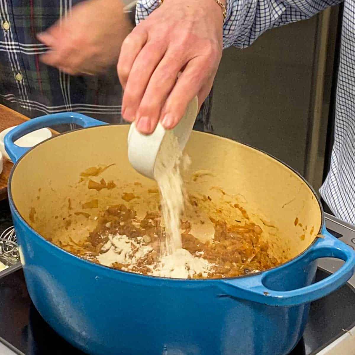 A person dumping all-purpose flour from a small bowl into a large Dutch oven that is filled with simmering caramelized onions.