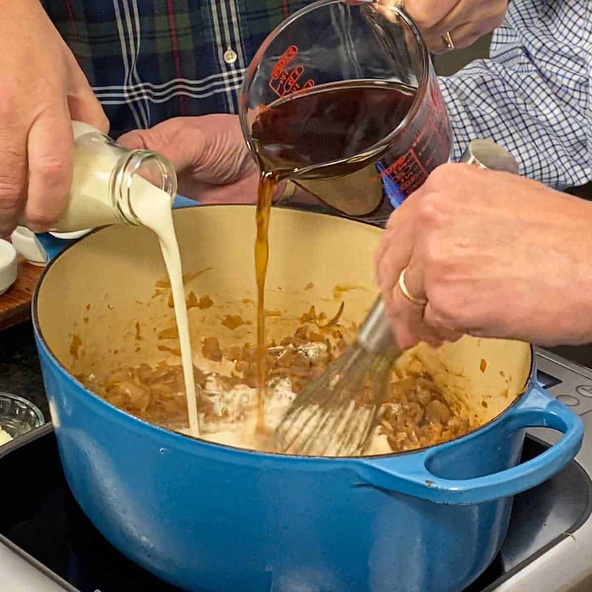 One person pouring heavy cream from a small milk jug and another person pouring beef stock from a measuring cup into a Dutch oven that is filled with flour-coated caramelized onions.