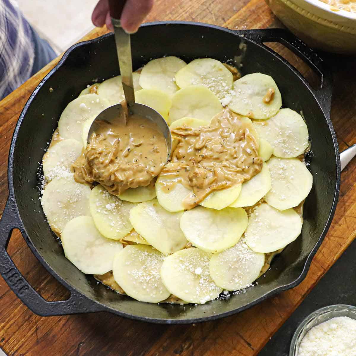 A person using a small ladle to transfer a French onion sauce over thinly sliced gold potatoe in a large cast-iron skillet.