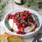 A circular dish holding Baked Brie with Cranberry-Orange Sauce and Pecans surrounded by two glasses of white wine, pieces of toast, holly, and silverware.