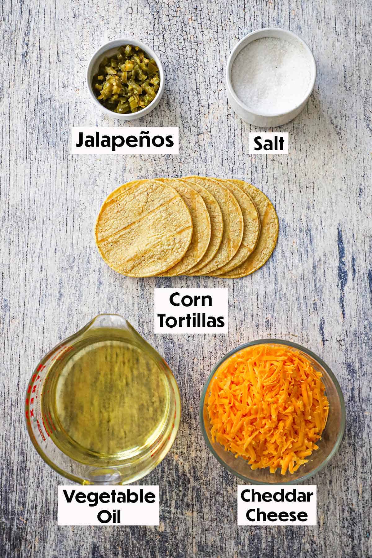 An arrangement of ingredients for classic nachos on a grey wooden background including corn tortillas, jalapeños, cheddar cheese, oil, and salt.