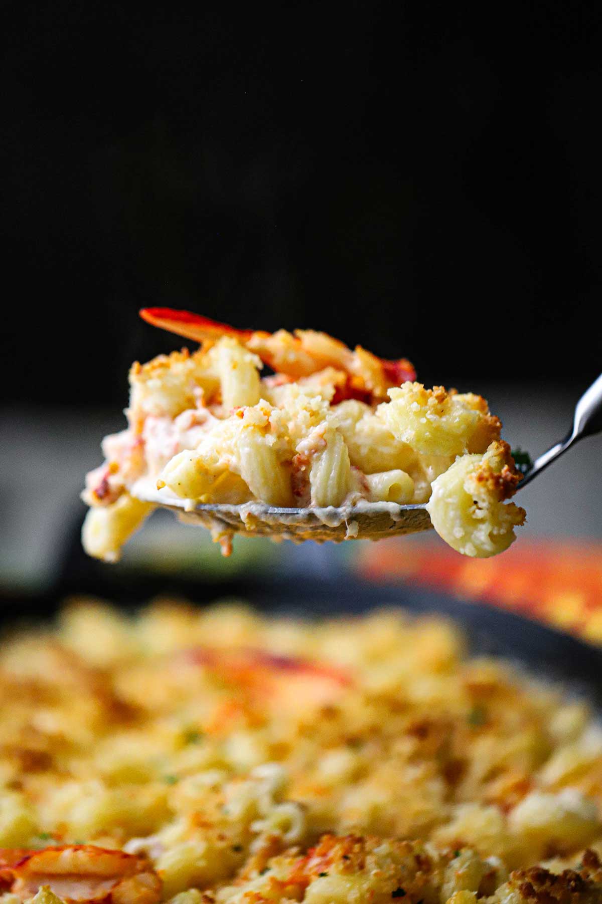 A large serving spoon being used to raise a serving of homemade lobster mac and cheese over a dish of the same.