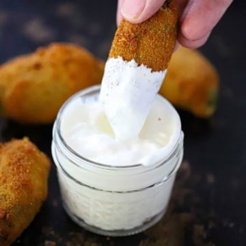 A person plunging a deep-fried jalapeño popper into a small glass jar filled with sour cream.