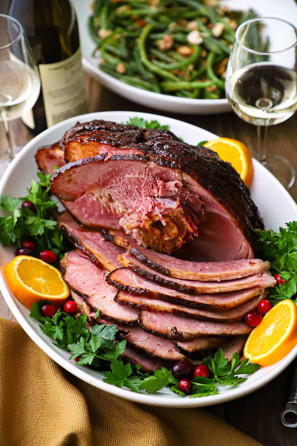 A large white platter filled with a honey mustard glazed smoked ham with two glasses of white wine and a bottle of wine nearby.