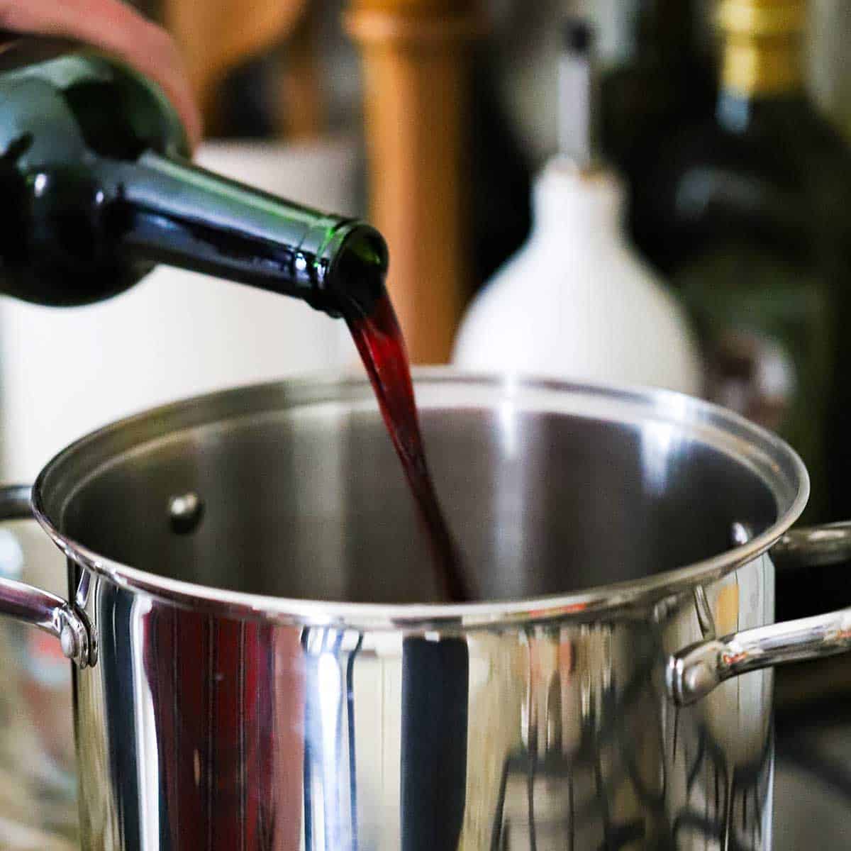 A person pouring red wine from a bottle into a silver stock pot on a gas stove.