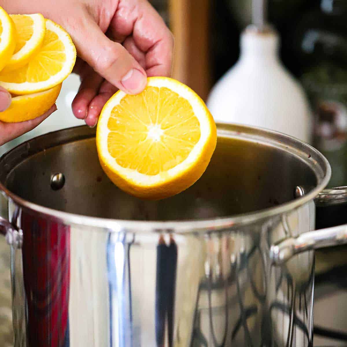 A person dropping slices of an orange into a silver stock pot on a gas stove.