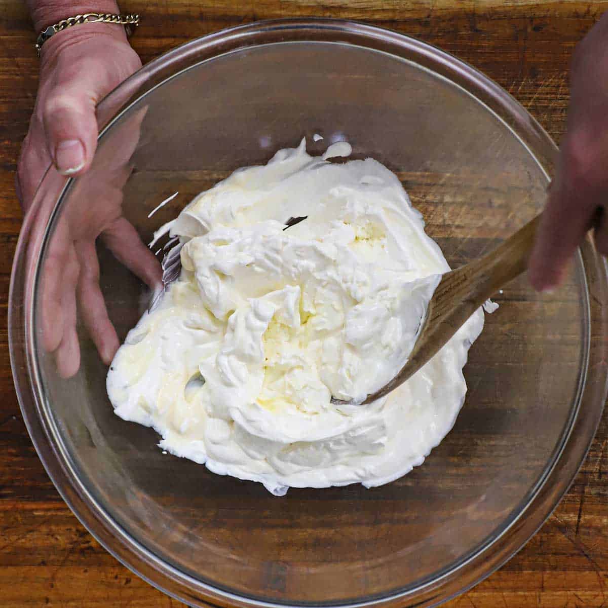 A person using a wooden spoon to mix softened cream cheese, sour cream, and mayonnaise in a glass bowl on a wooden cutting board.