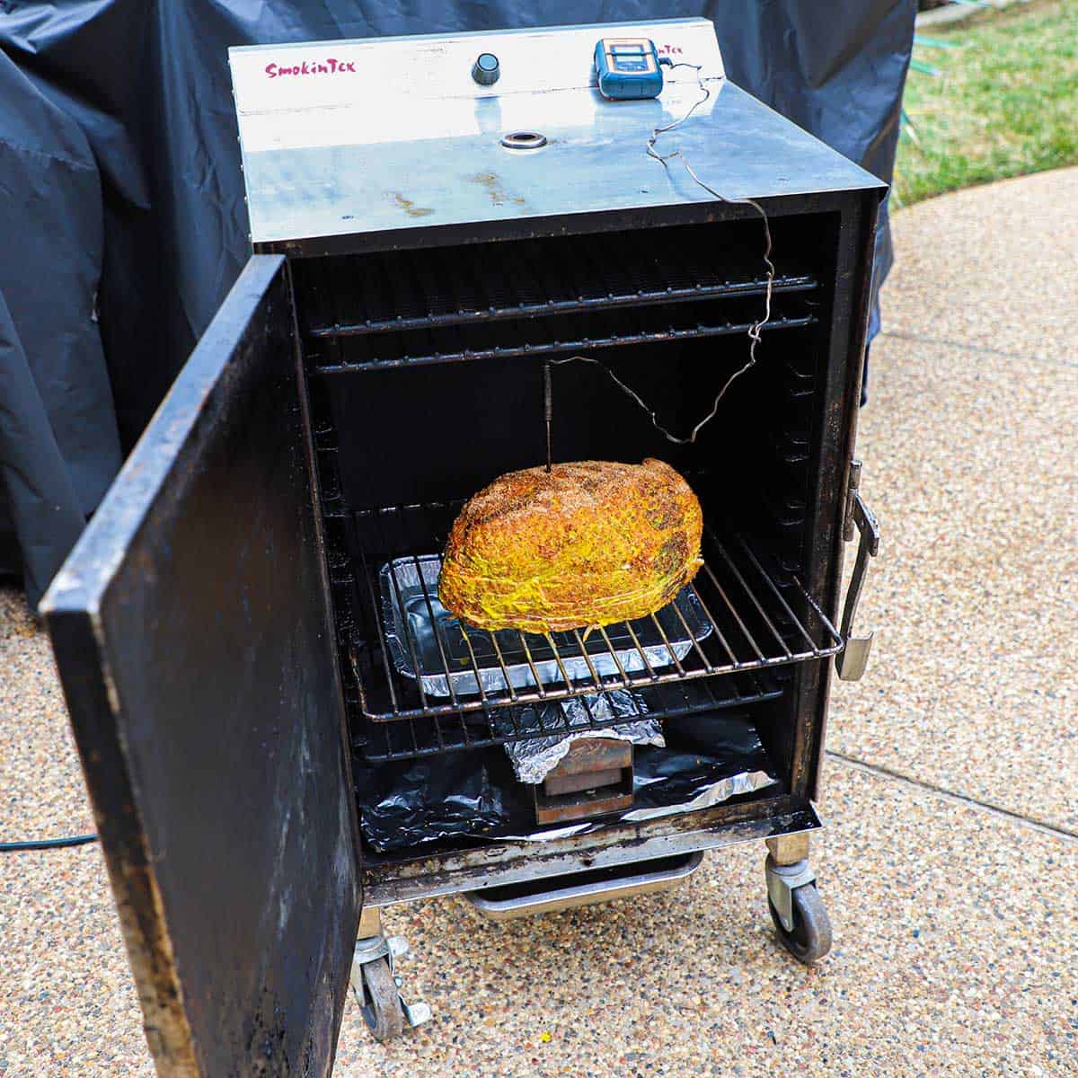 An eight pound Berkshire spiral-cut ham that has been coated with yellow mustard and pork rub sitting inside an electric smoker with a digital thermometer stuck in the middle and the door of the smoker open.