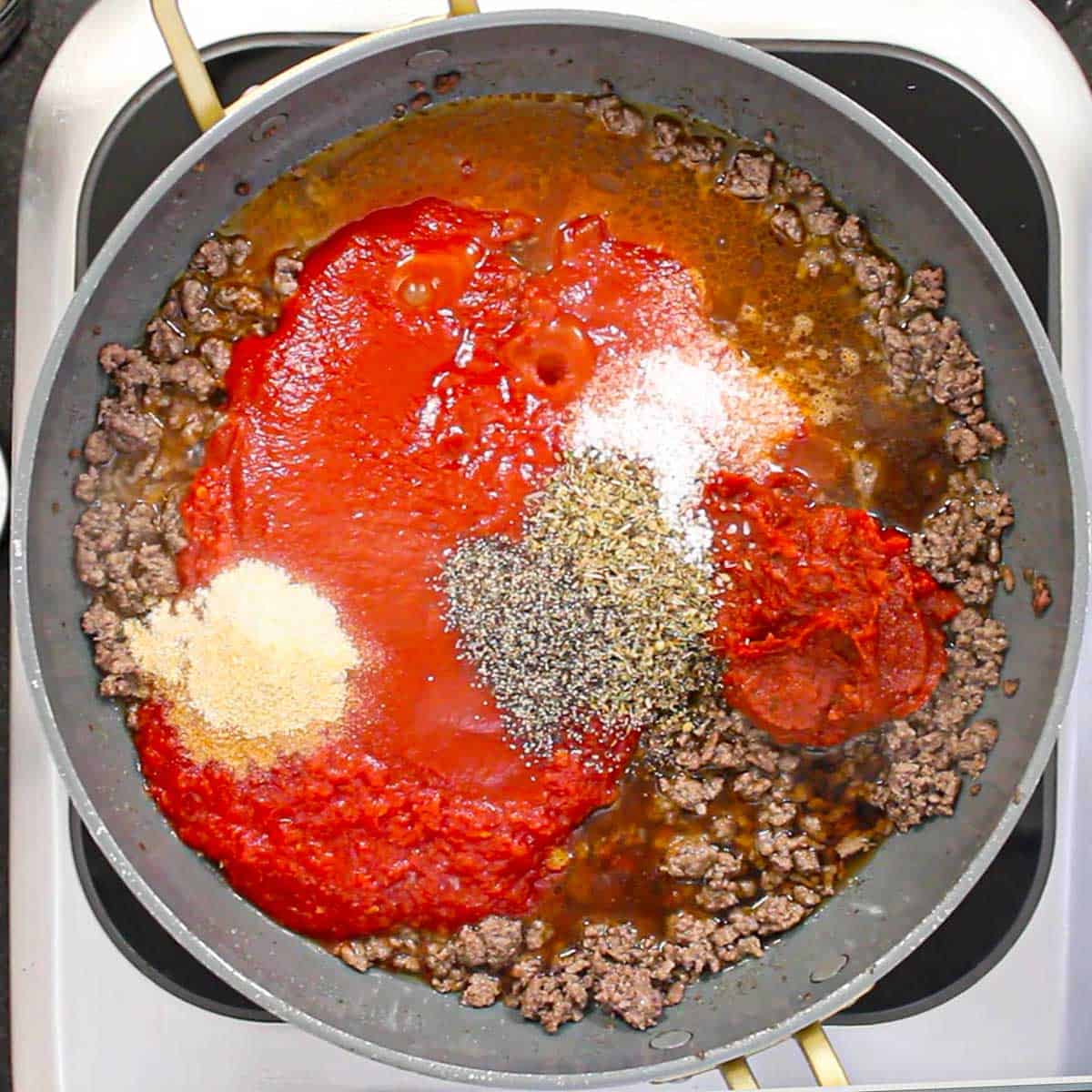 A large non-stick skillet filled with cooked ground beef, tomato sauce, beef stock, tomato paste, and seasonings.