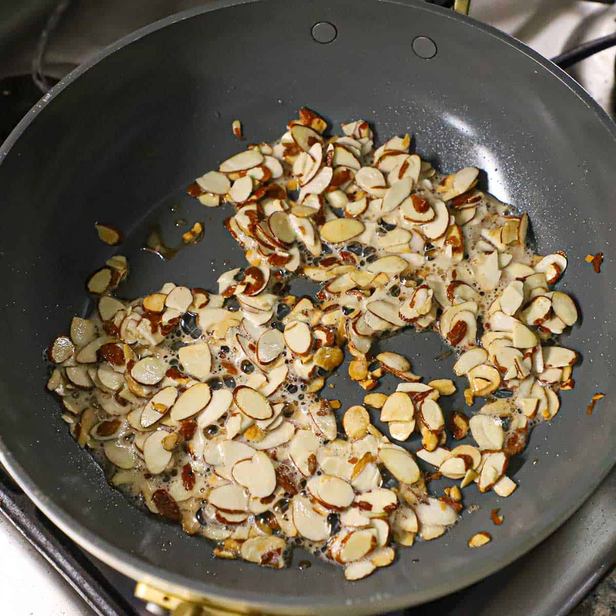 A large non-stick skillet filled with sliced almonds being lightly browned in oil and melted butter on a gas stove.