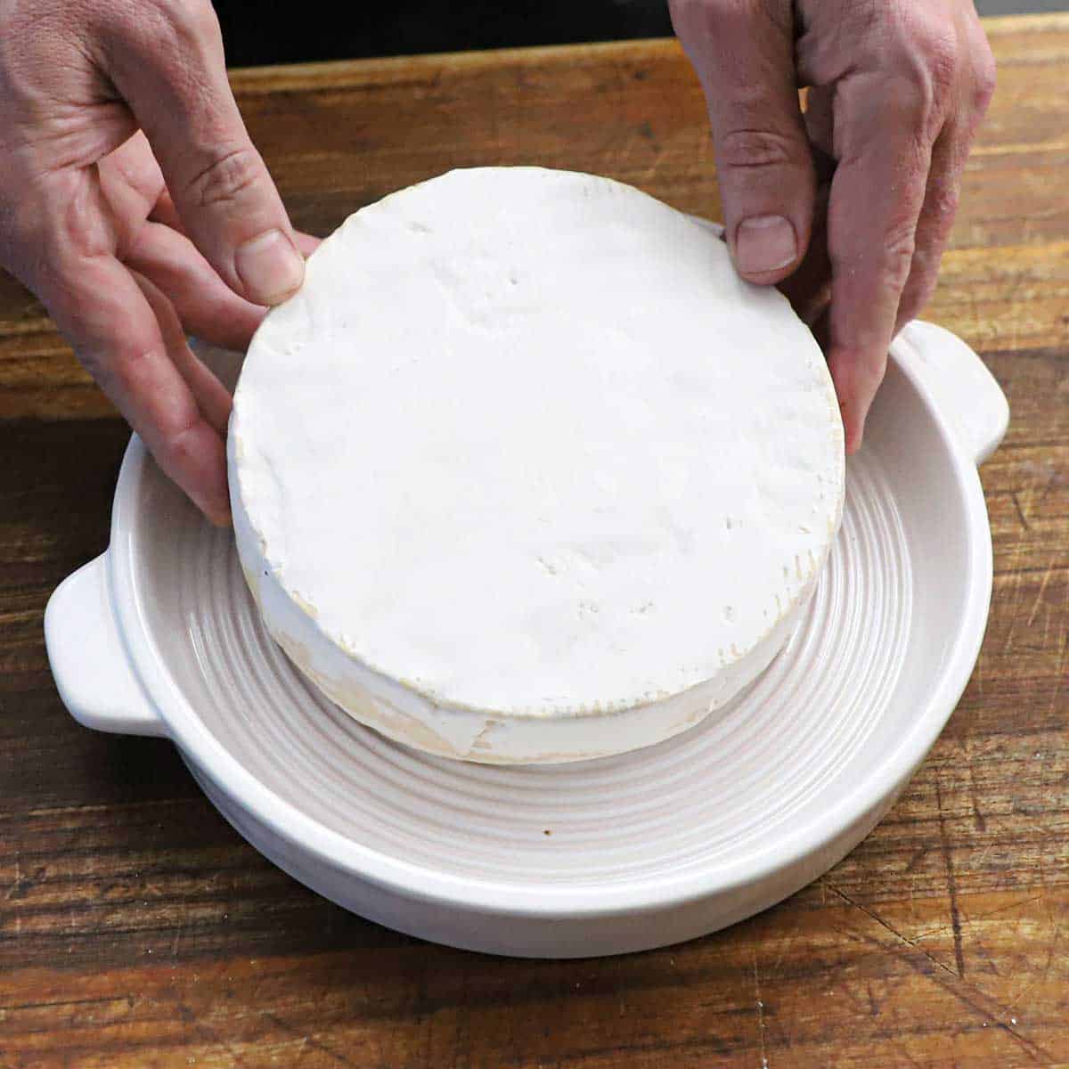 A person placing a wheel of brie into a circular baking dish on a wooden cutting board.