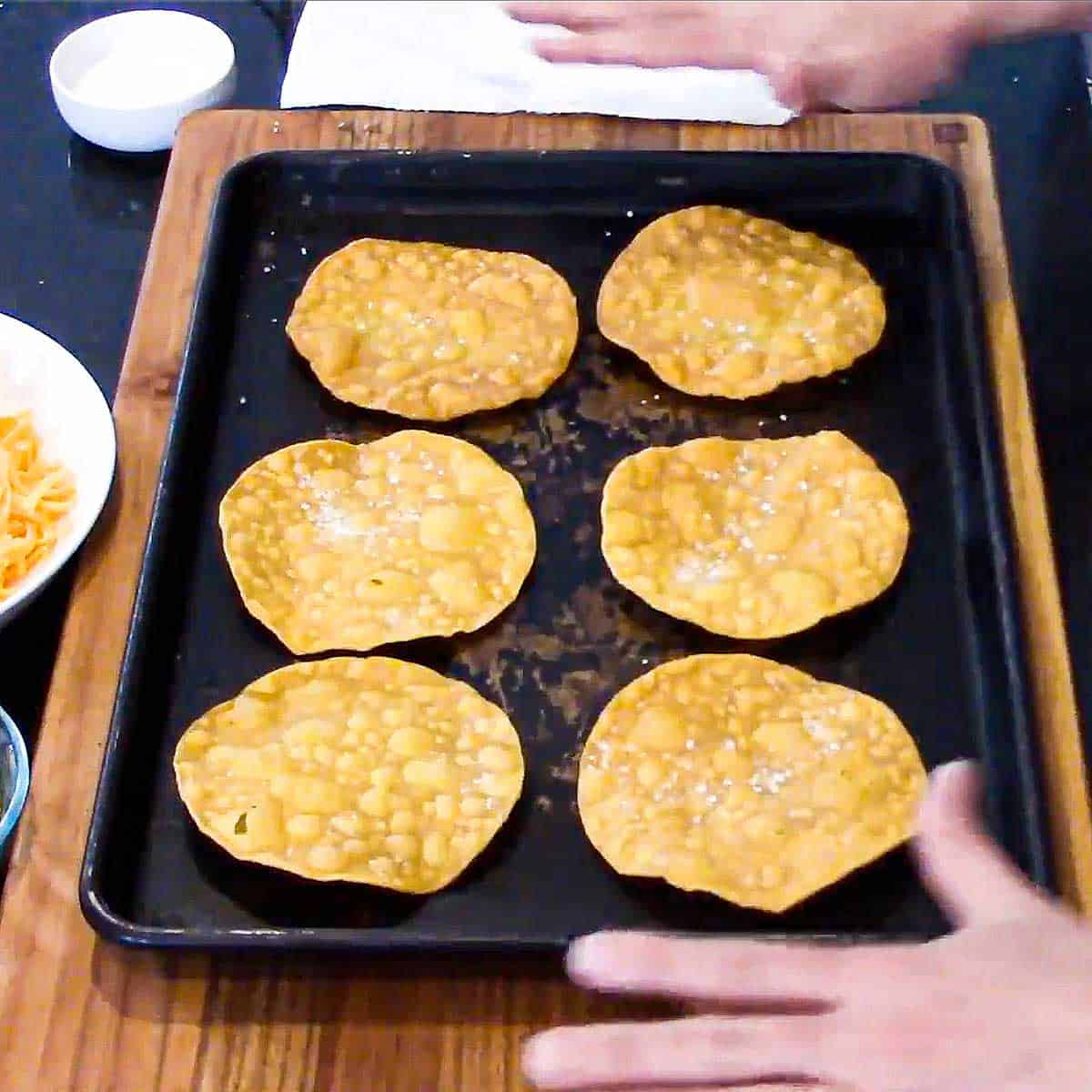 A person standing near a baking sheet that is holding six deep-fried corn tortillas that have been lightly salted.