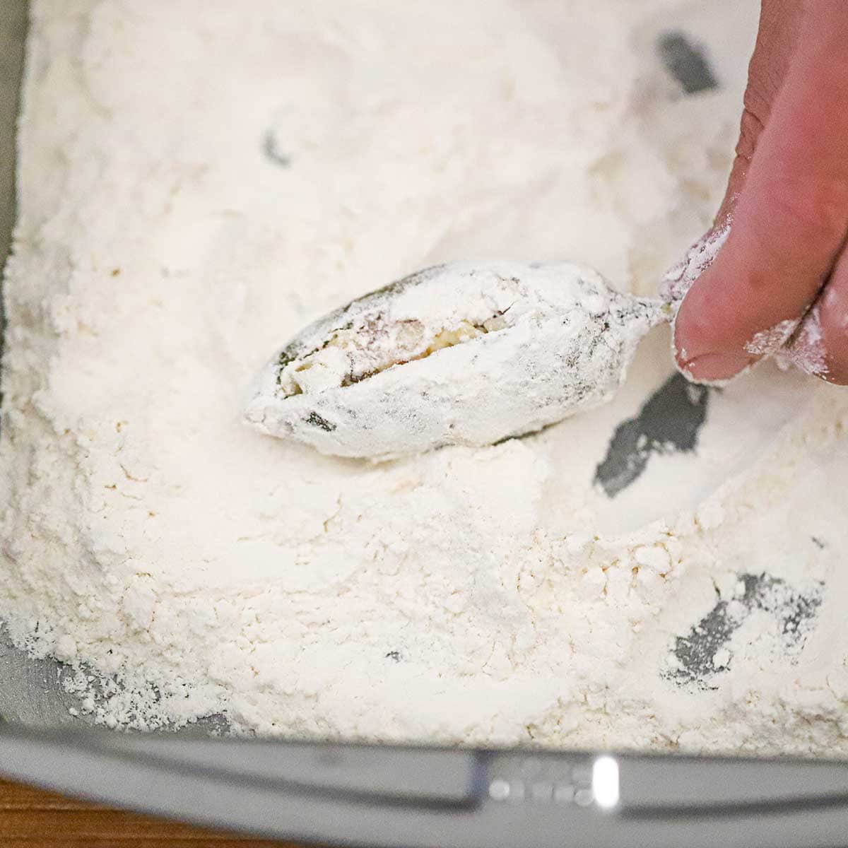 A person dredging a stuffed jalapeño through a pan filled with flour.