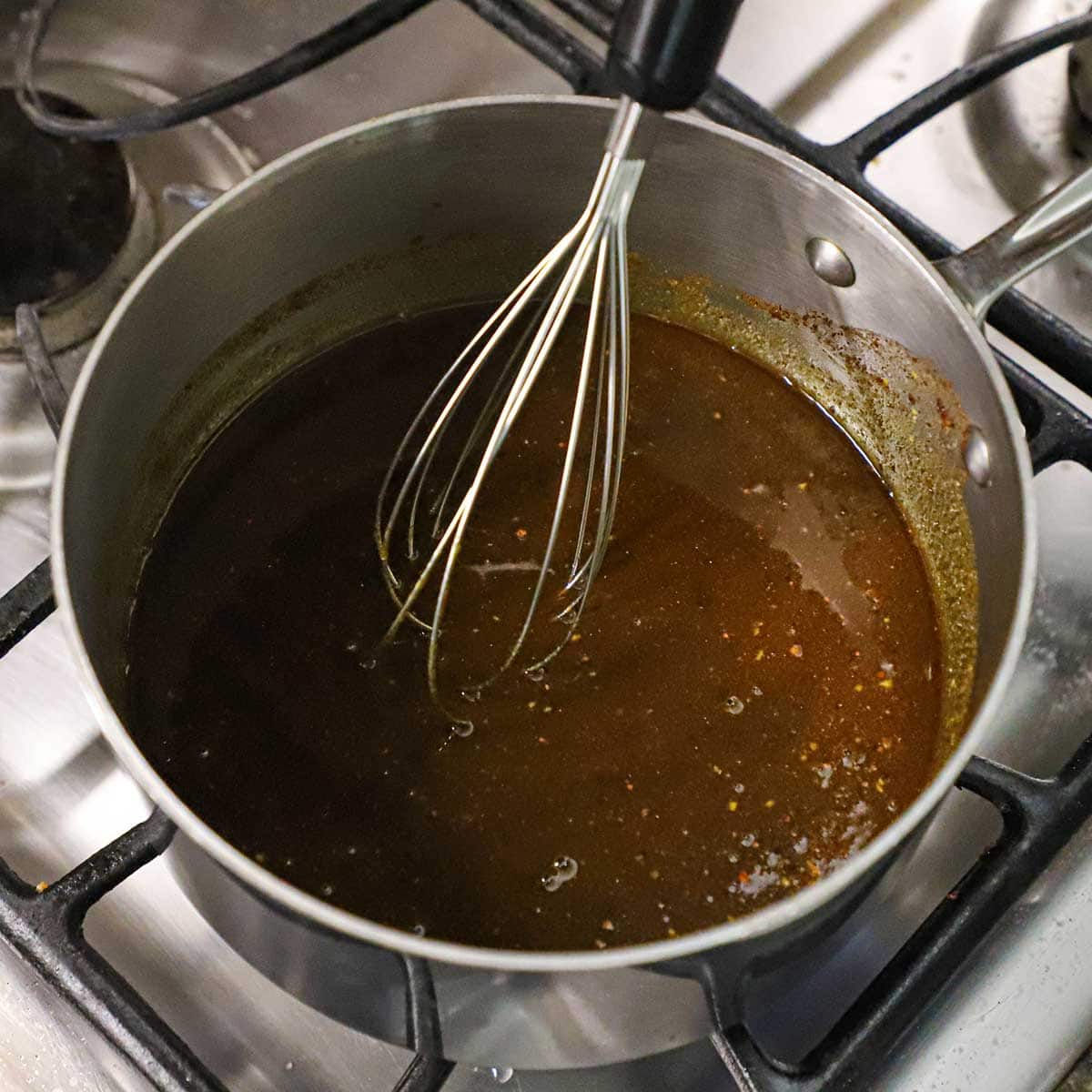 A person using a metal whisk to stir a honey mustard glaze for ham in a saucepan on a gas stove.