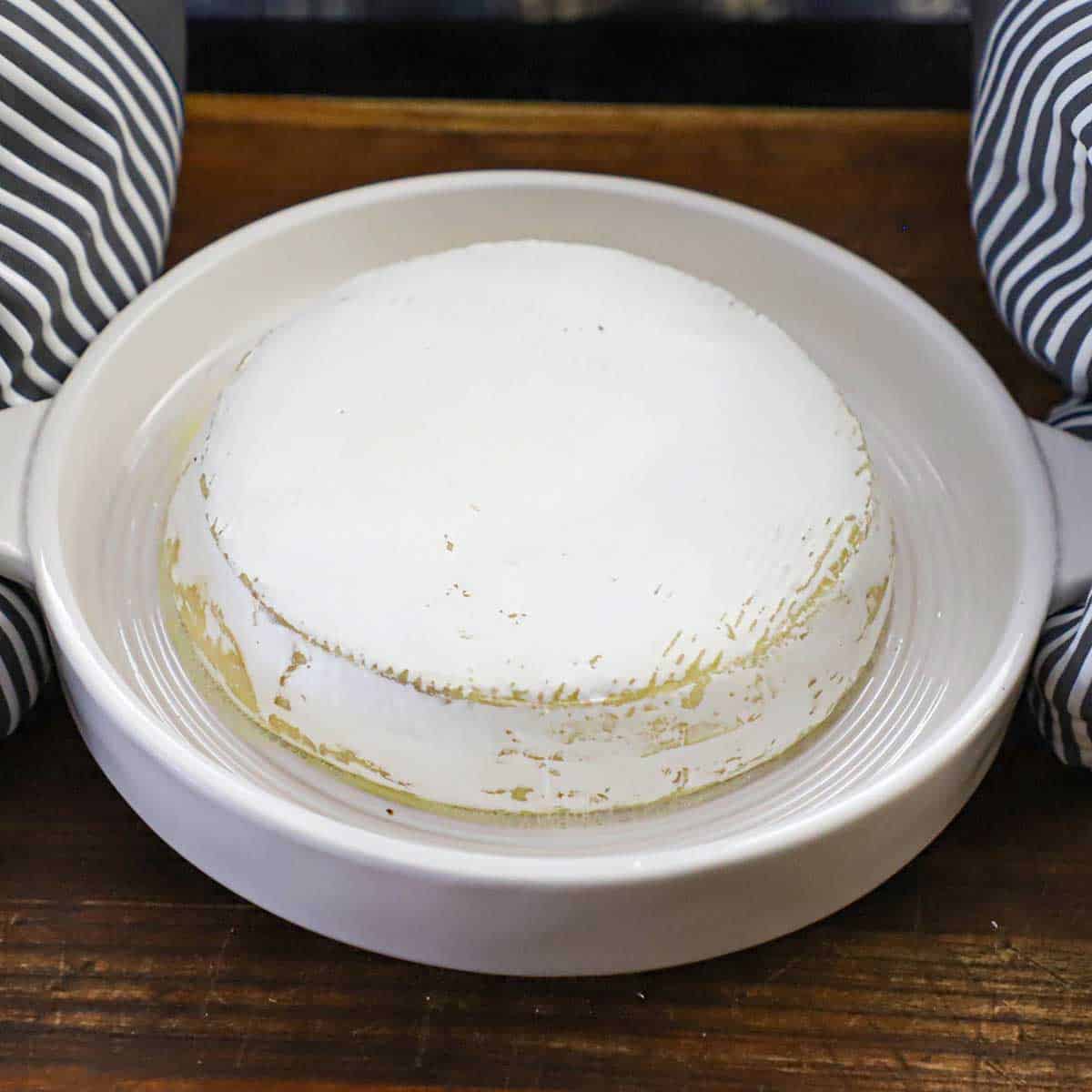 A person where oven mitts holding the handles of a circular baking dish that is holding a wheel of brie that has been heated in the oven.