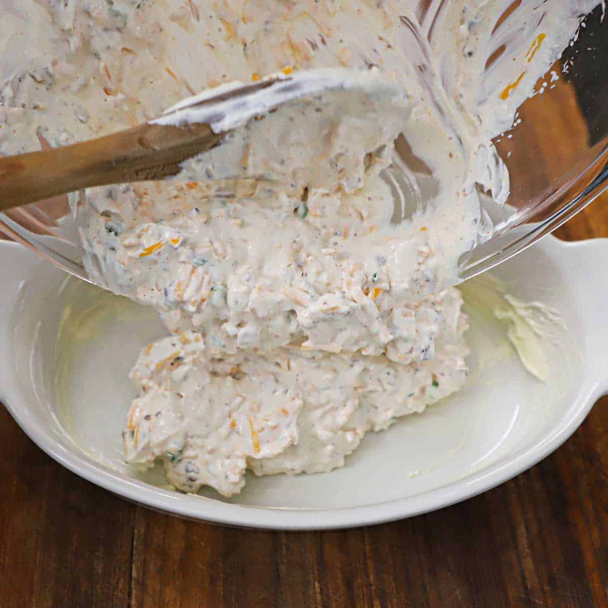 A person using a large wooden spoon to transfer uncooked bacon and cheddar dip from a large glass bowl into a greased oval baking dish.
