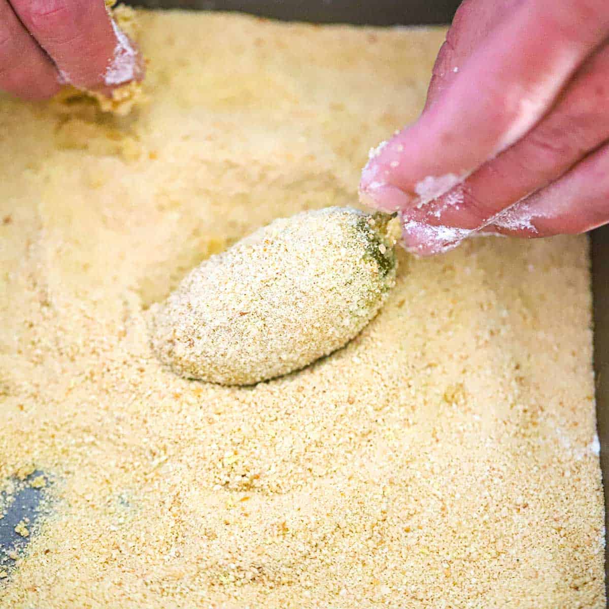 A person dredging an uncooked jalapeño popper through a pan that is filled with fine breadcrumbs.