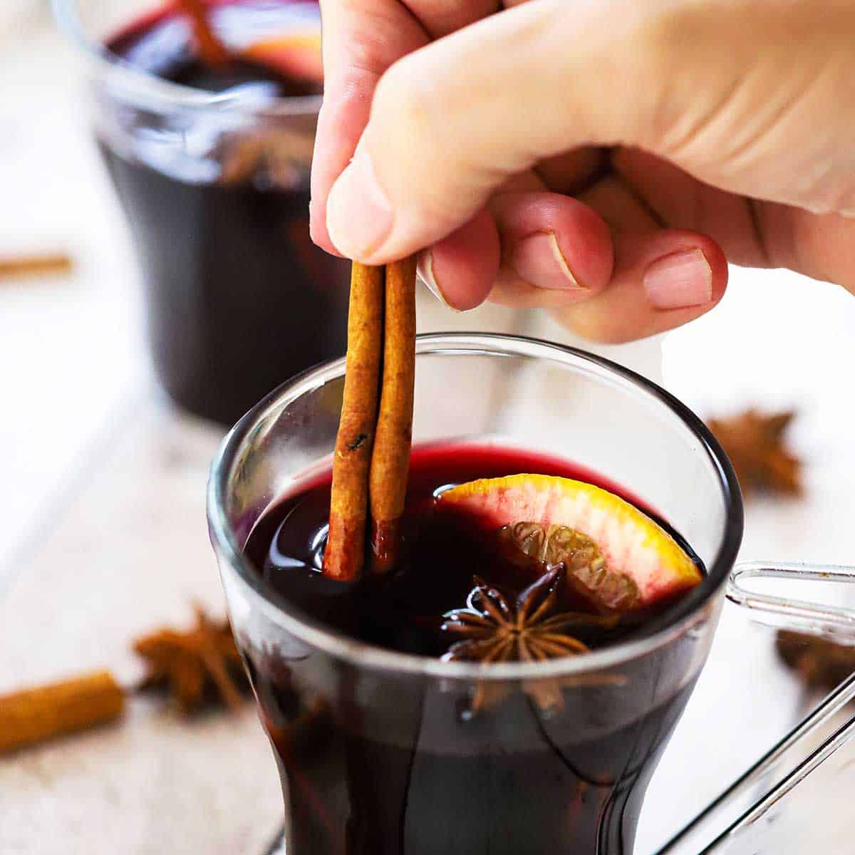 A person placing two cinnamon stick into a glass mug that is filled with glehwein (mulled wine).