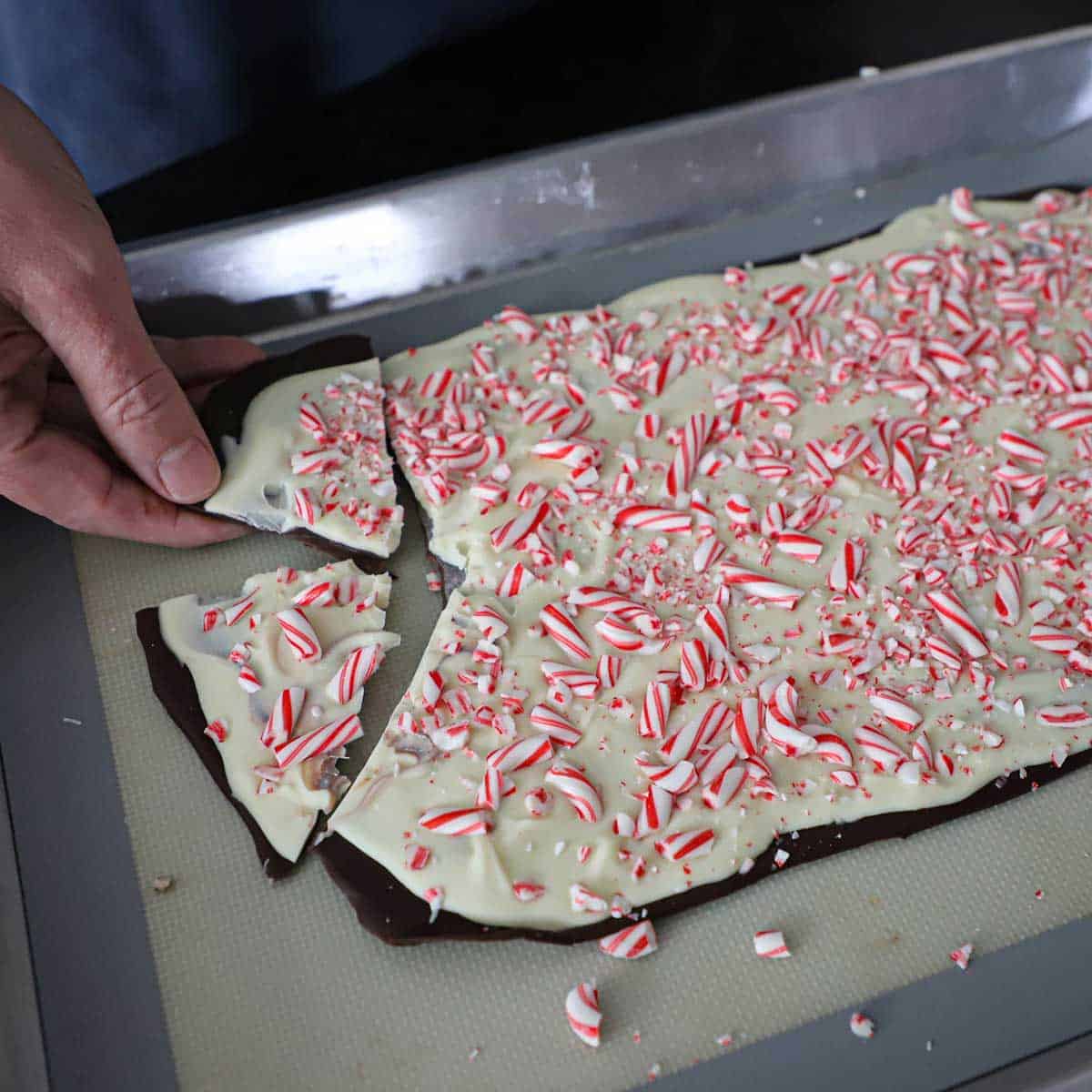 A person using his hand to break pieces of chilled homemade peppermint bark from a large block of it that is resting in a half sheet pan on a silicone matt.