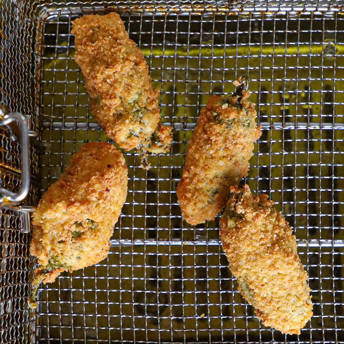 Four homemade jalapeño poppers that are resting in a fry basket above the oil in a deep-fryer.