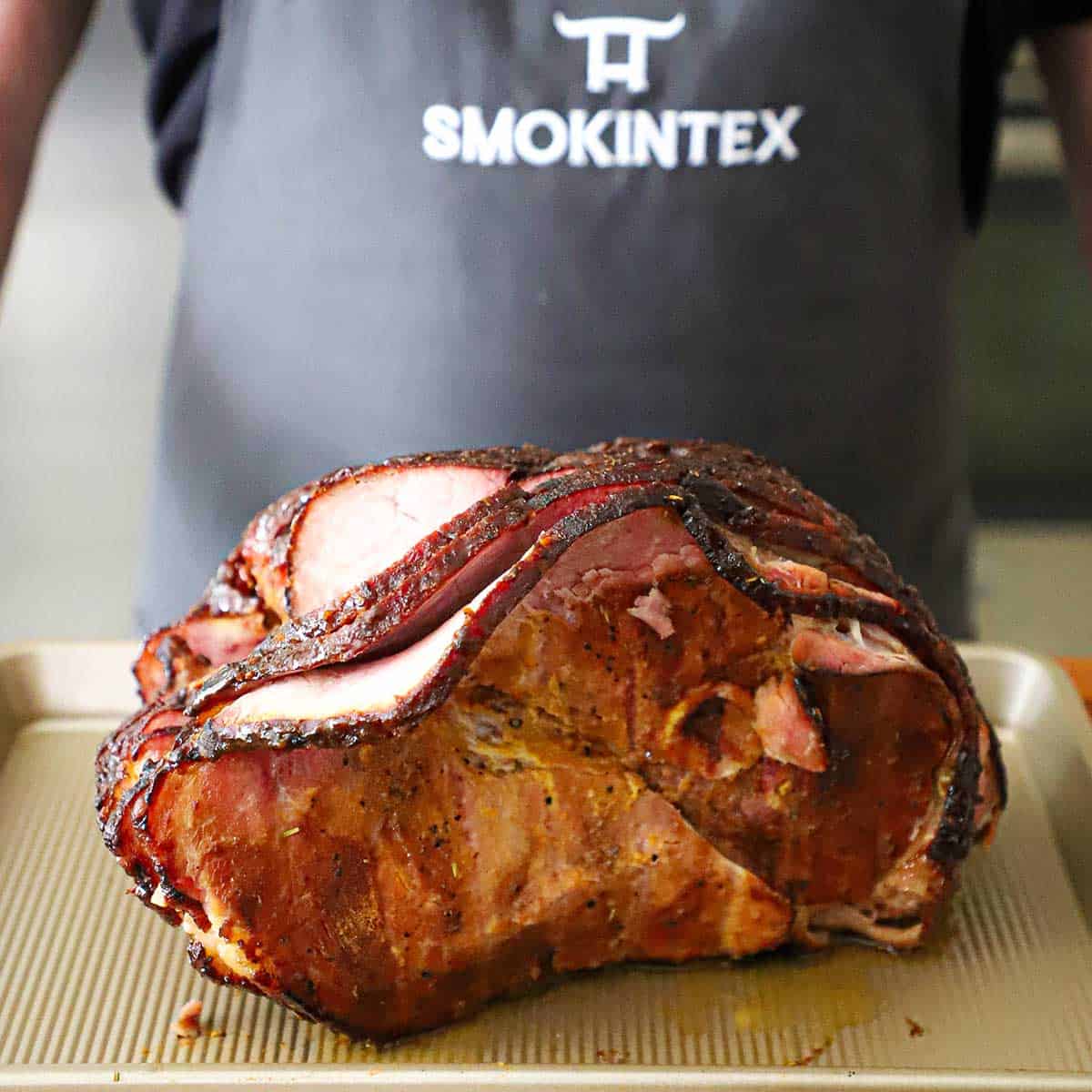 A person wearing a Smokin' Tex apron standing behind a honey mustard smoked ham that is resting on a sheet.