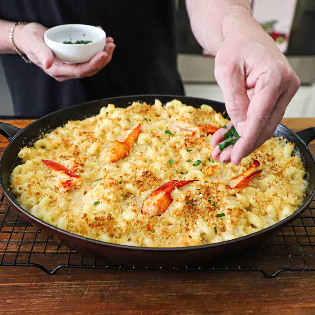 A person sprinkling chopped Italian parsley over the top of a freshly baked homemade lobster mac and cheese in an oval baking dish.